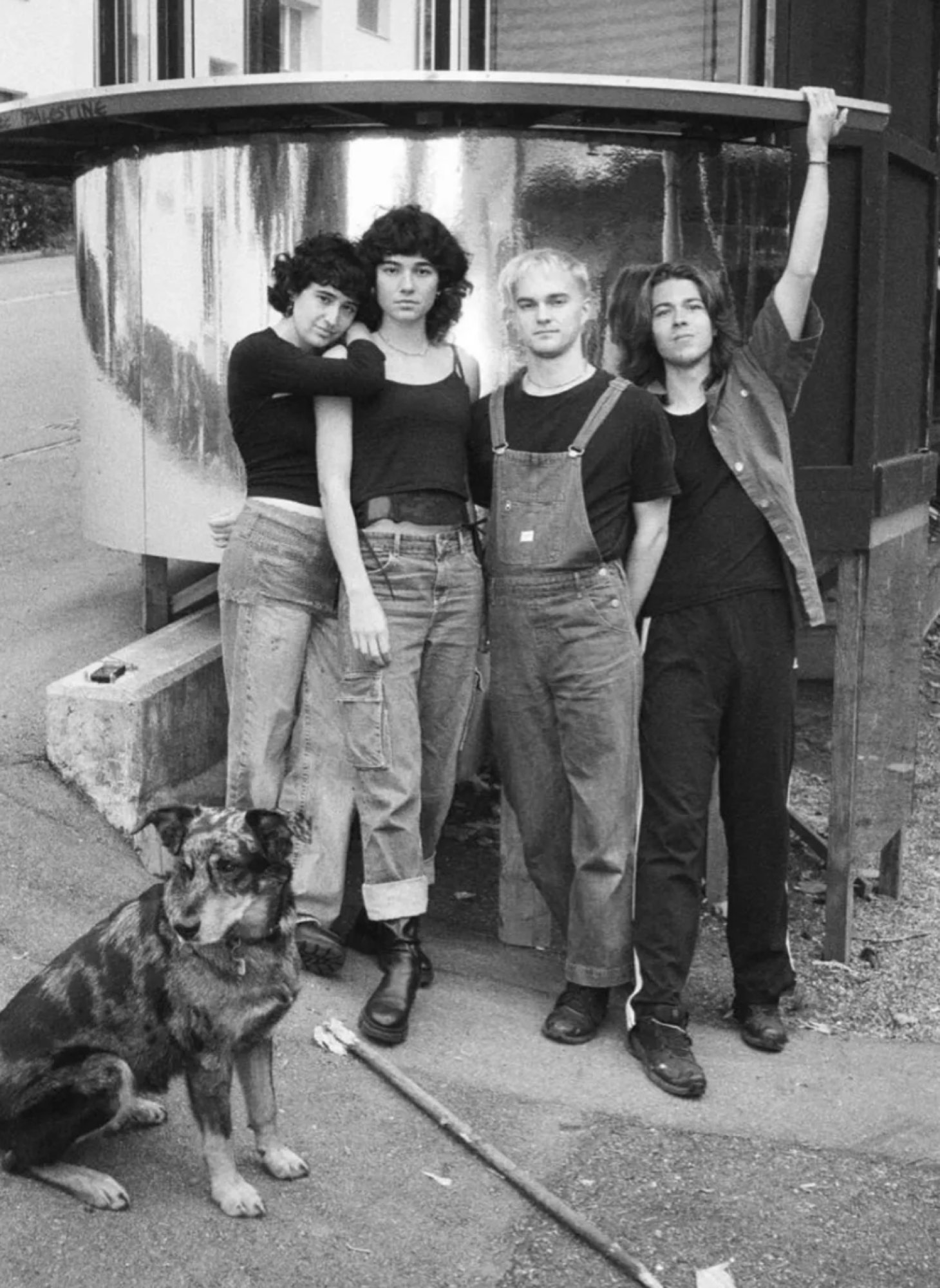 Band members of Deluxe Lynx including singer-songwriter Camille Santacroce. Image Description: Black and white photo of four young people standing outdoors in front of an agricultural barn in Switzerland, with a dog sitting nearby.