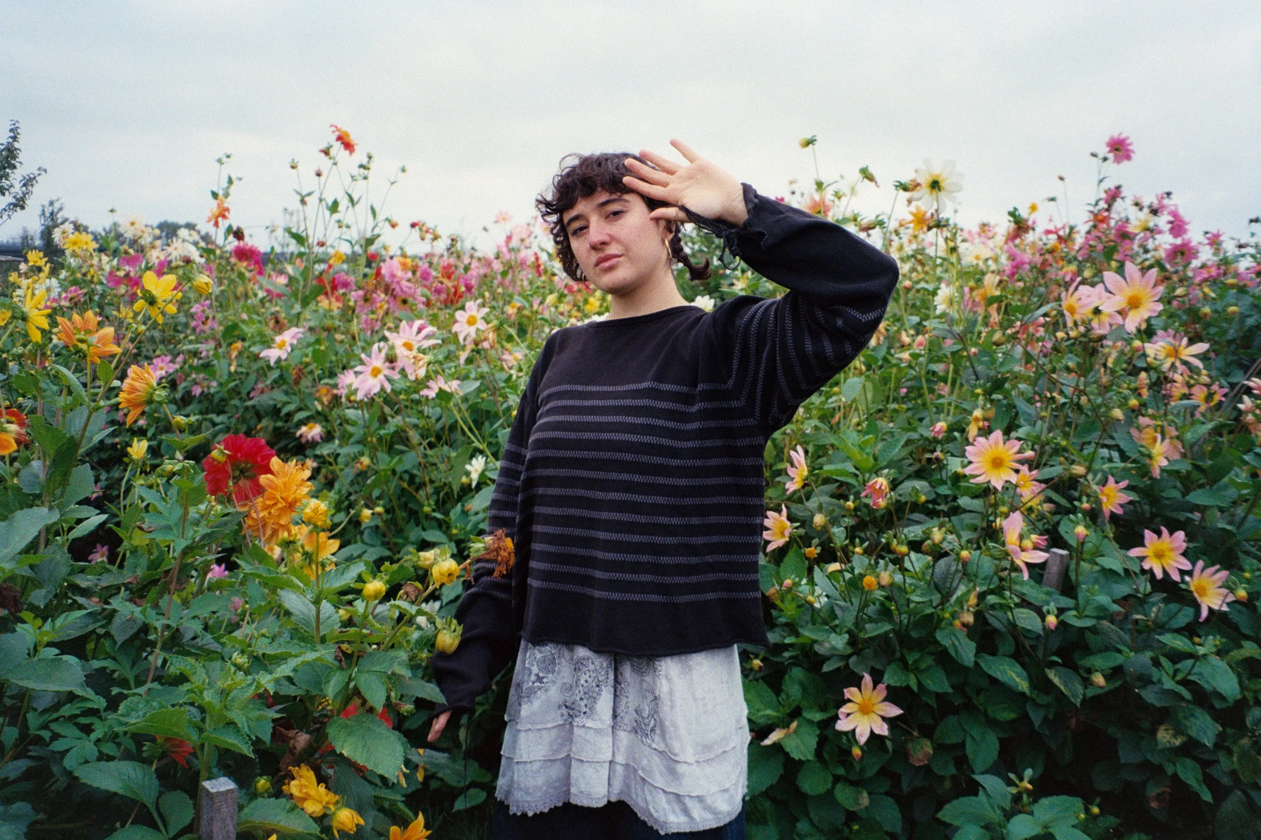 Swiss singer-songwriter Camille Santacroce standing in a colorful flower garden, wearing a black striped sweater and a layered white skirt, with her right hand raised near their head.
