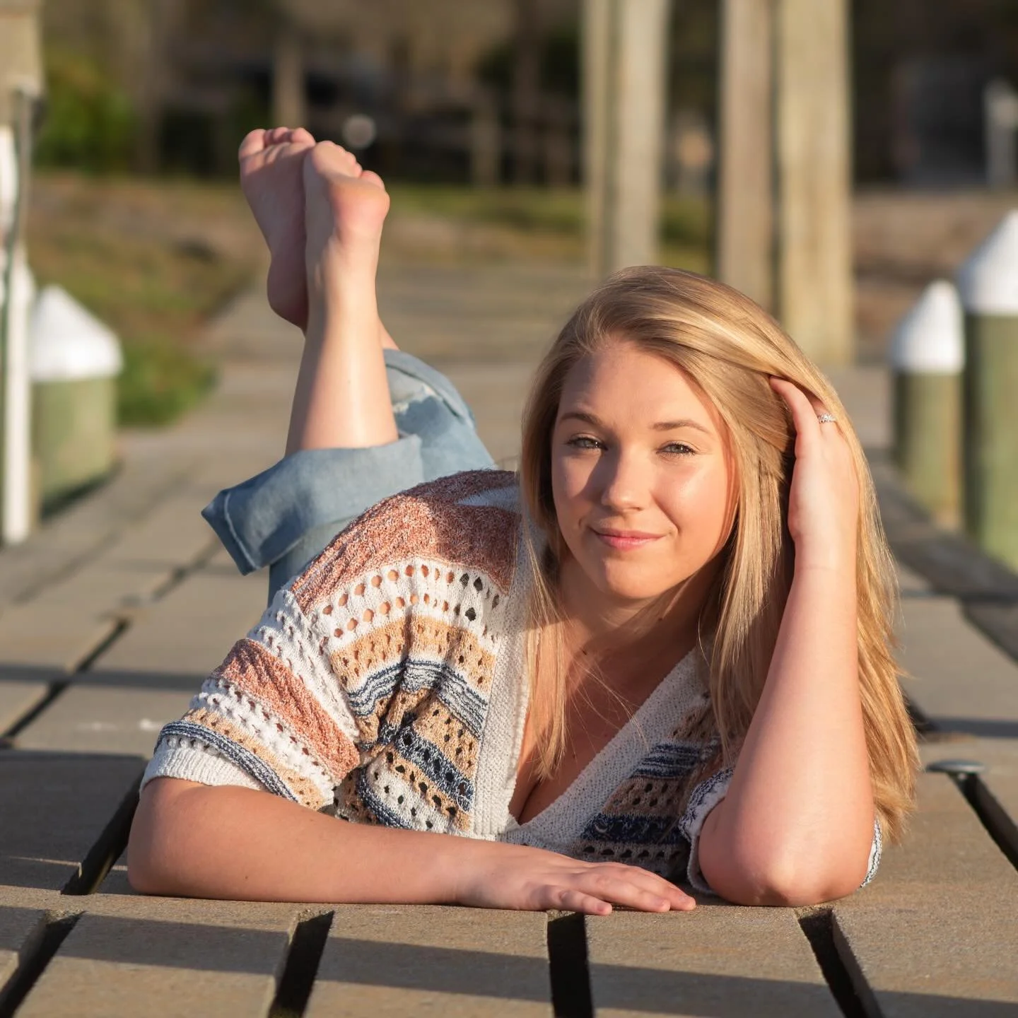A young woman with blonde hair lying on a wooden dock, smiling, with one hand resting on her head and her legs crossed in the air.