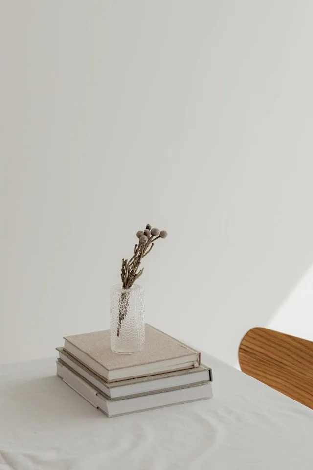 A clear textured glass vase with dried gray flower stems on top of four stacked books with neutral-colored covers, placed on a white table near a wooden chair.