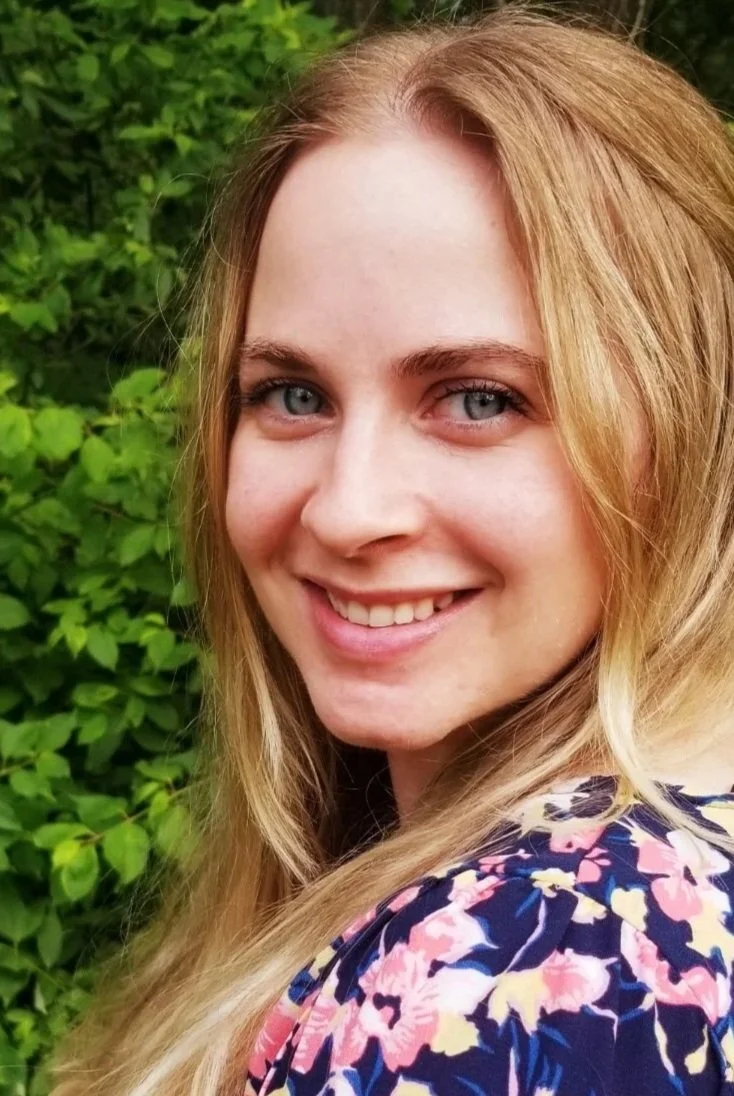 Close-up of a smiling young woman with blonde hair and blue eyes, standing outdoors in front of green foliage.