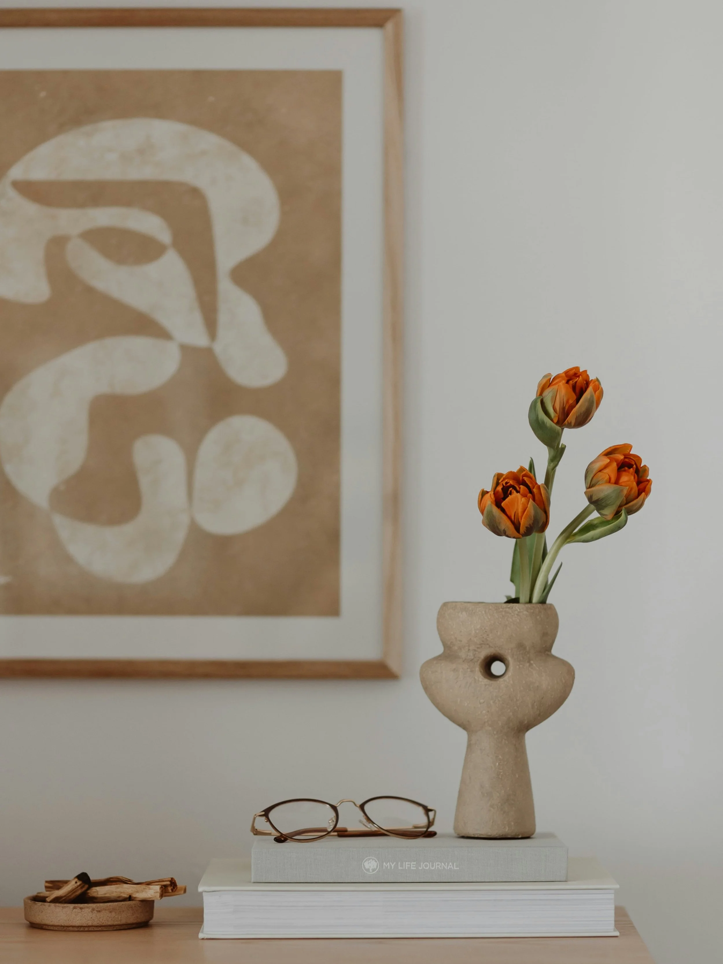 Minimalist beige ceramic vase with orange and green flowers on top of a stack of books, with a pair of eyeglasses on the bottom book, and artwork in the background.