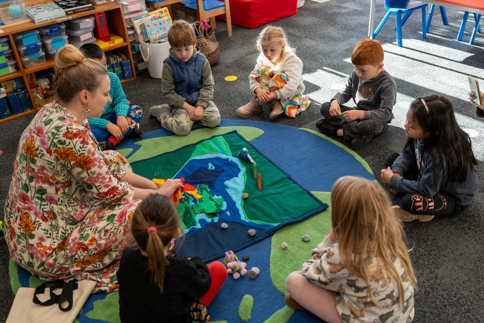 Children sitting in a circle on a colorful rug, playing with a fabric topographical map of Greenland and surrounding areas, inside a classroom.