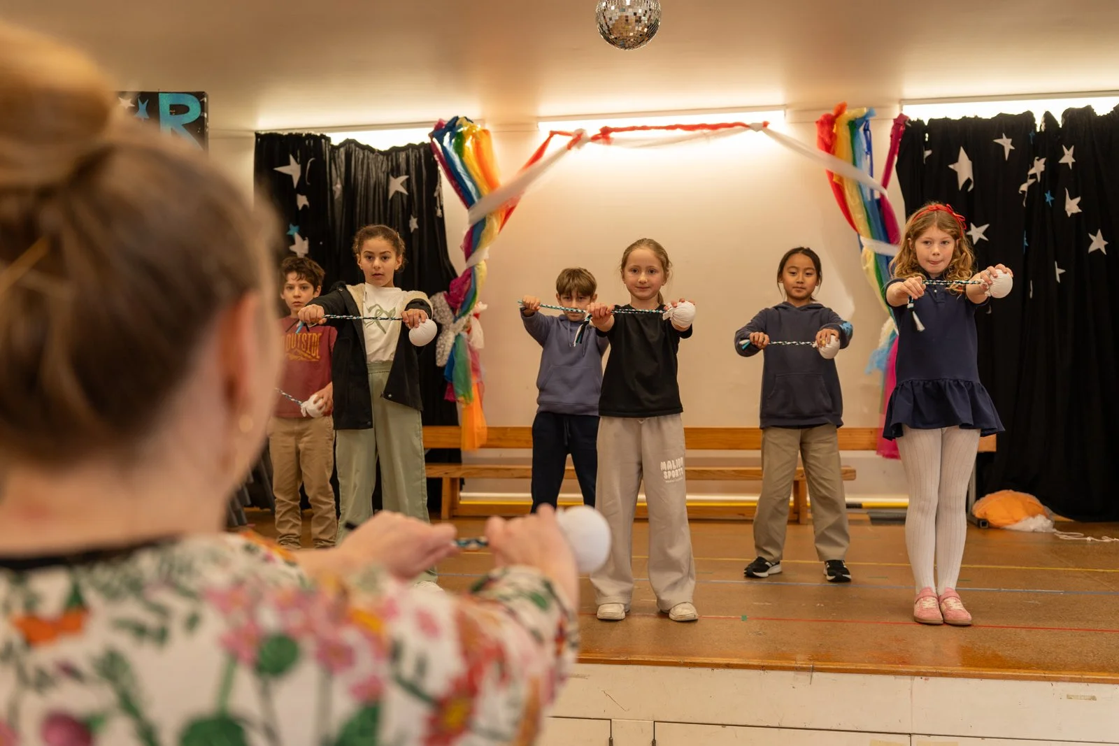 Children performing a choreographed dance on stage during a school event, with a woman in floral clothing watching from the foreground.