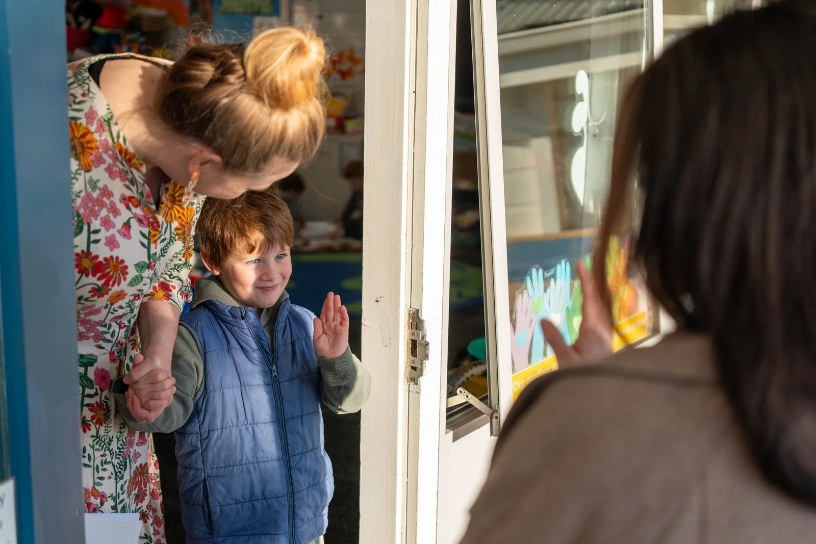 A young boy standing at a colorful toy store entrance, waving and smiling at a woman inside, with a woman and a store employee visible in the background.
