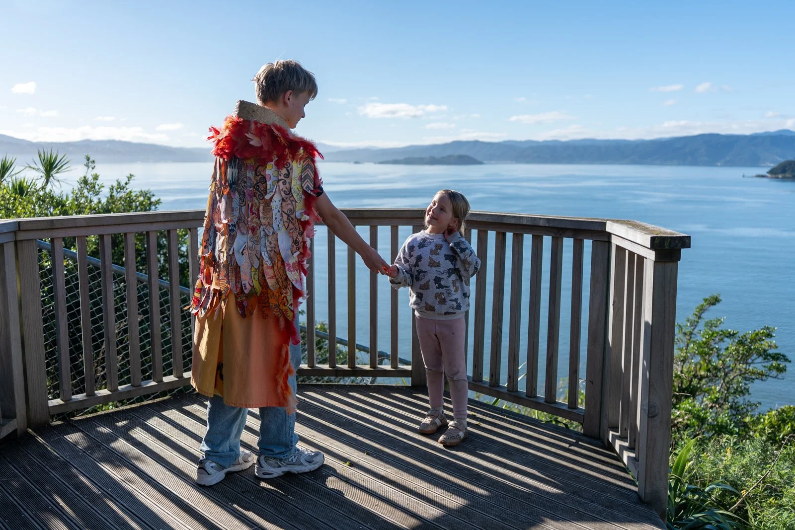 A woman and a young girl standing on a wooden balcony overlooking a large body of water with mountains in the background, holding hands.
