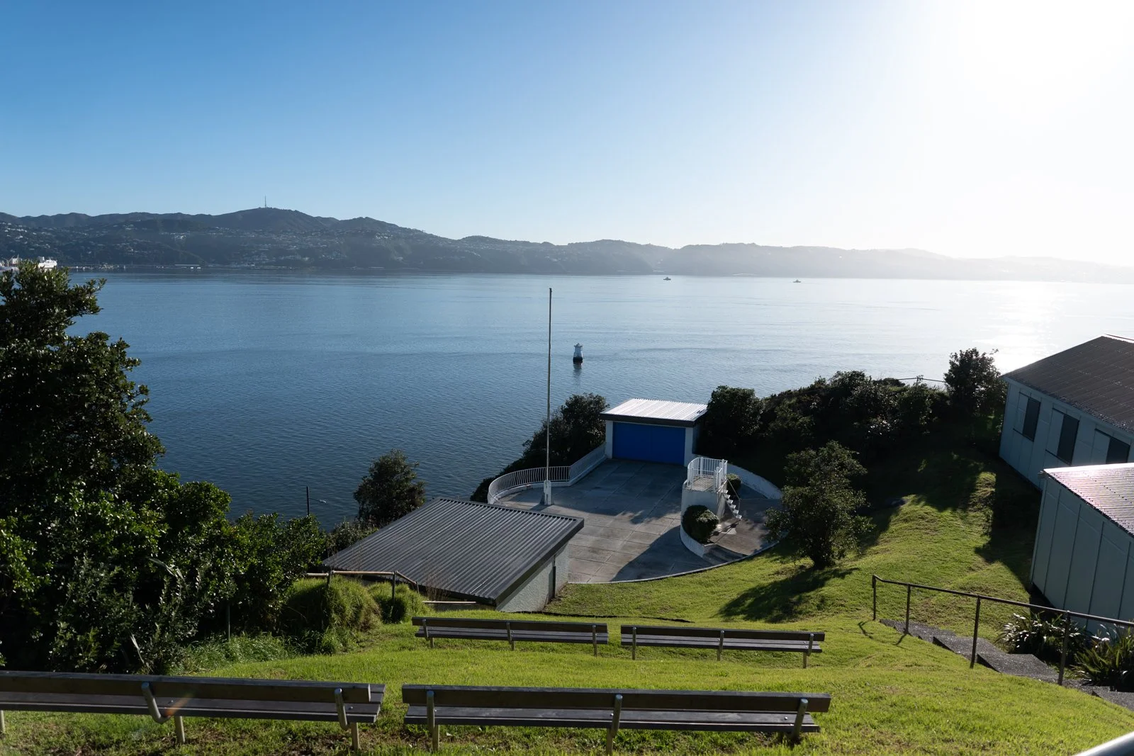 A peaceful waterfront view showing a calm body of water with hills in the background, a small dock with a buoy in the water, and a grassy area with trees, buildings, a concrete patio, and benches in the foreground.