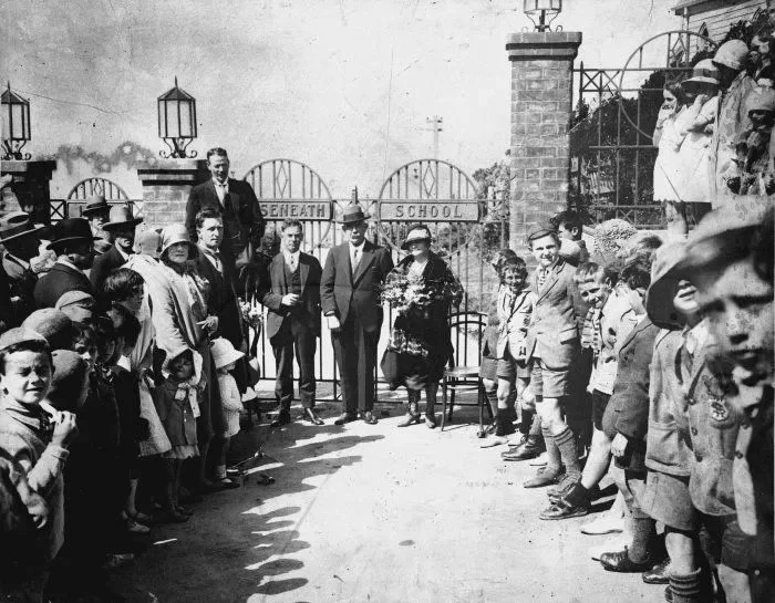 A group of children and adults standing outdoors in a semicircle for a school event outside a gate with a sign that reads "SENEATH SCHOOL." Several adults are at the center, with one holding a bouquet of flowers. Children are dressed in vintage clothing, and some are wearing hats.