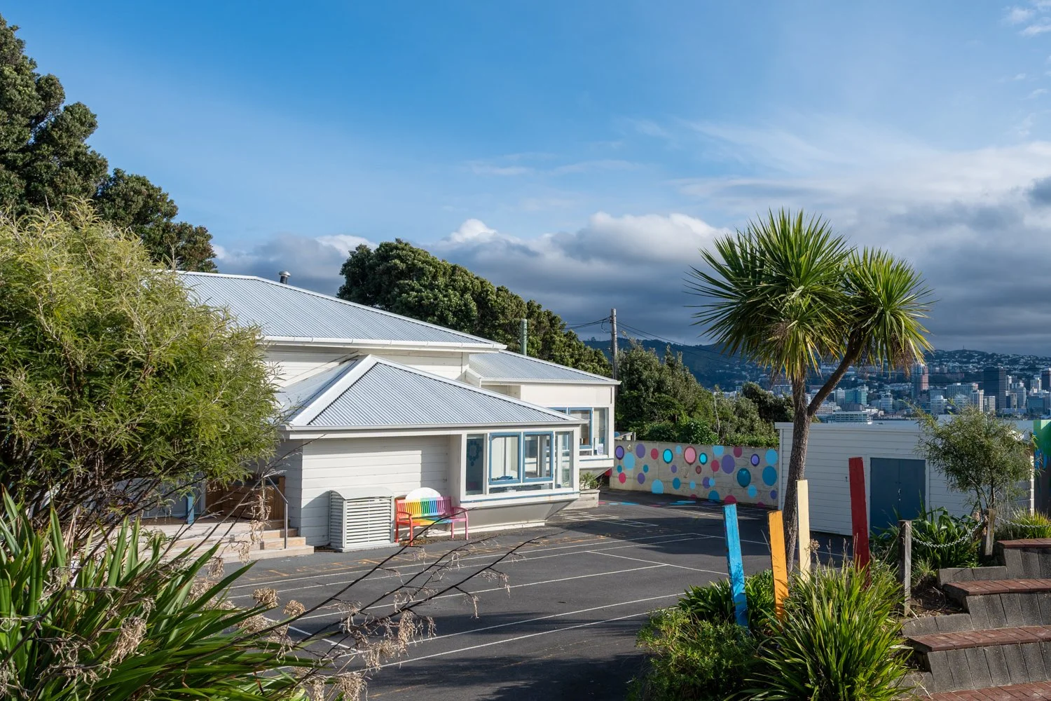 Sunny day view of white residential buildings, a parking lot, colorful painted poles, a palm tree, lush greenery, and cityscape in the background.