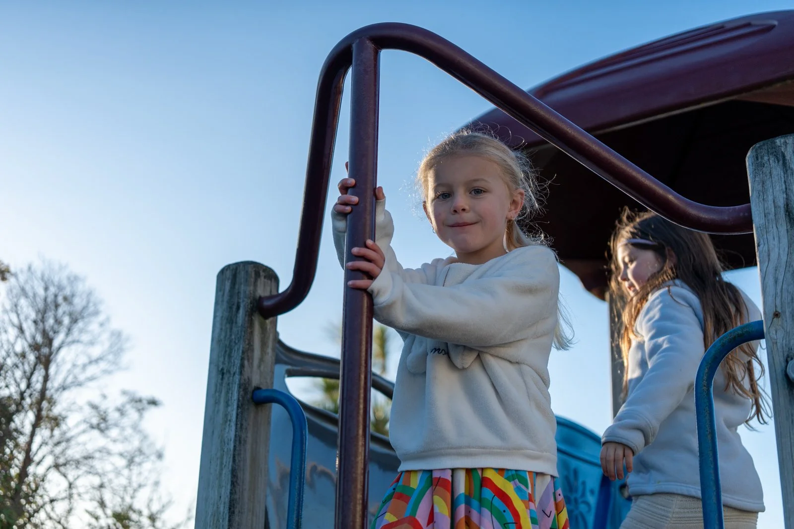 Two young girls playing on a playground slide, one girl is holding the railing and smiling at the camera, while the other girl is standing behind her, looking down.
