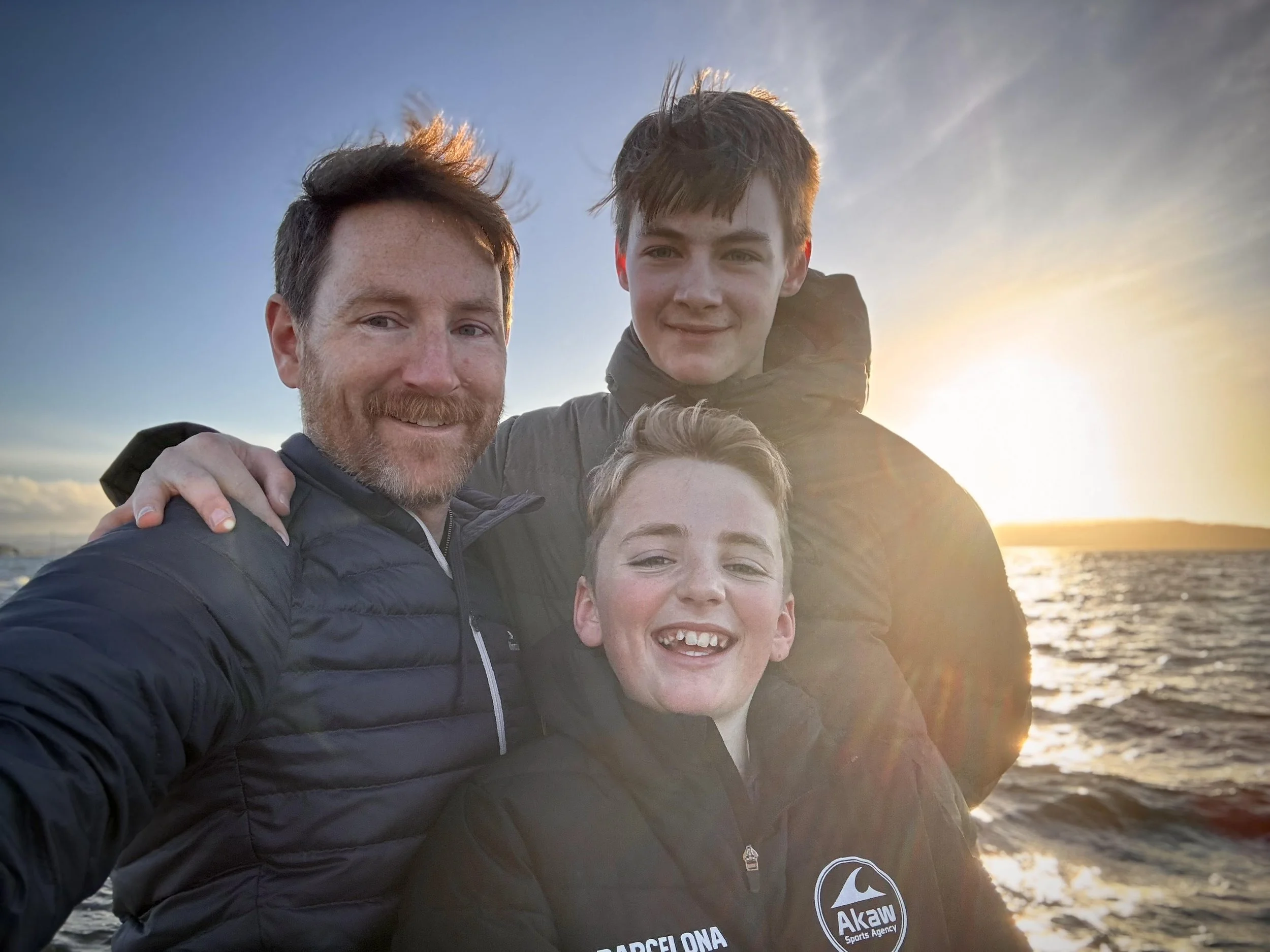 Four people smiling at the camera during sunset at the beach, with a body of water and a cloudy sky in the background.