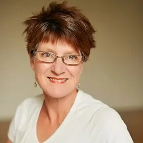 A woman with short, spiky reddish-brown hair, glasses, and a white top smiling at the camera.