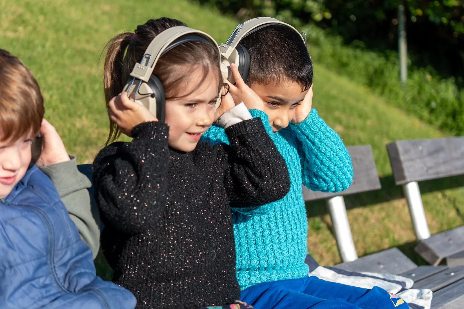 Three children sitting on a park bench, enjoying music on headphones.