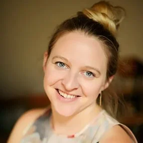 A young woman with blonde hair tied in a bun, smiling, wearing a sleeveless top.