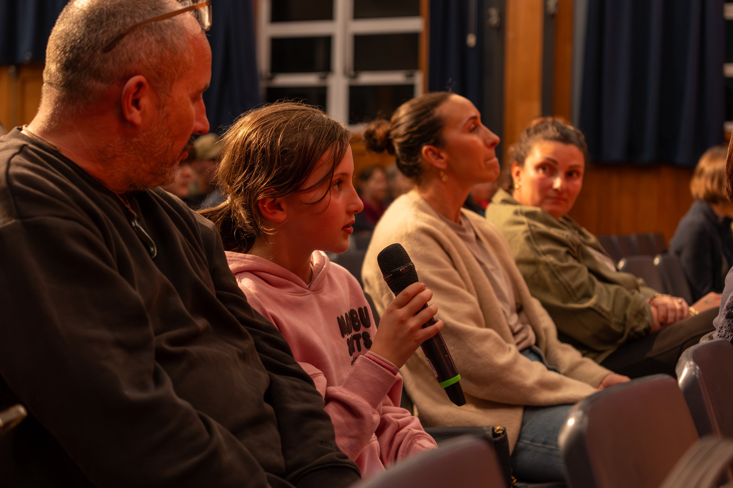 Group of people sitting in a row at an indoor event, with one woman holding a microphone.