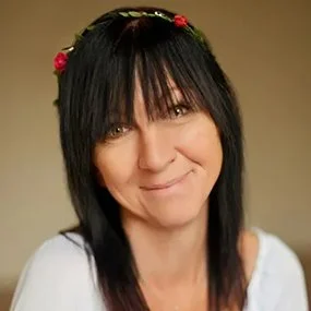 A woman with black hair and bangs, wearing a flower crown, smiling at the camera.