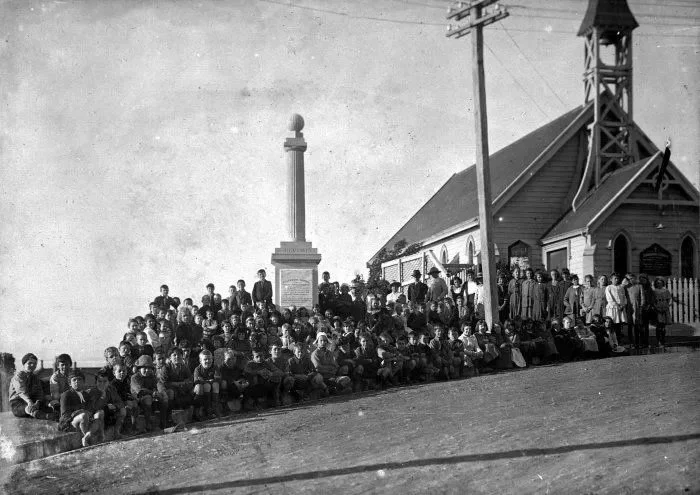 Black-and-white photo of a large group of children and a few adults gathered outside a church with a tower, monument, and utility poles nearby.