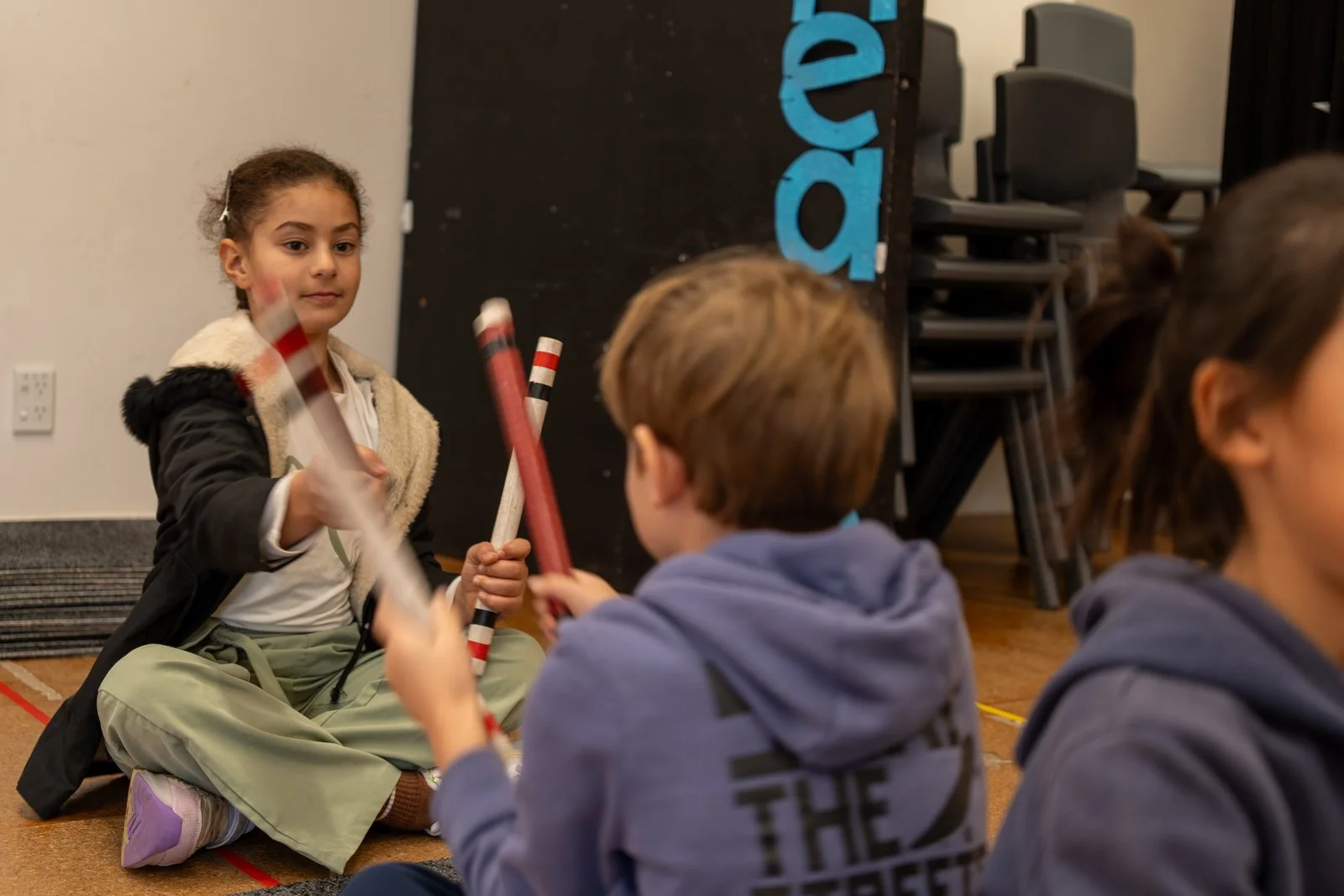 Children sitting on the floor in a circle, engaging in a musical activity with rhythm sticks, in a classroom or indoor setting.