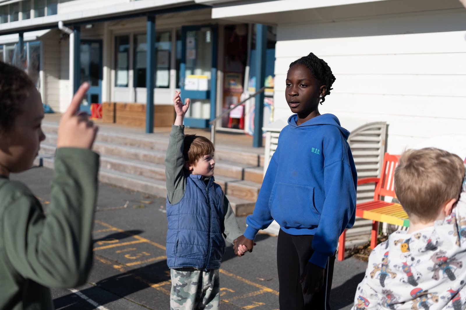 Children standing outside a building, two children holding hands and others raising hands, with a white wall, stairs, and benches in the background.