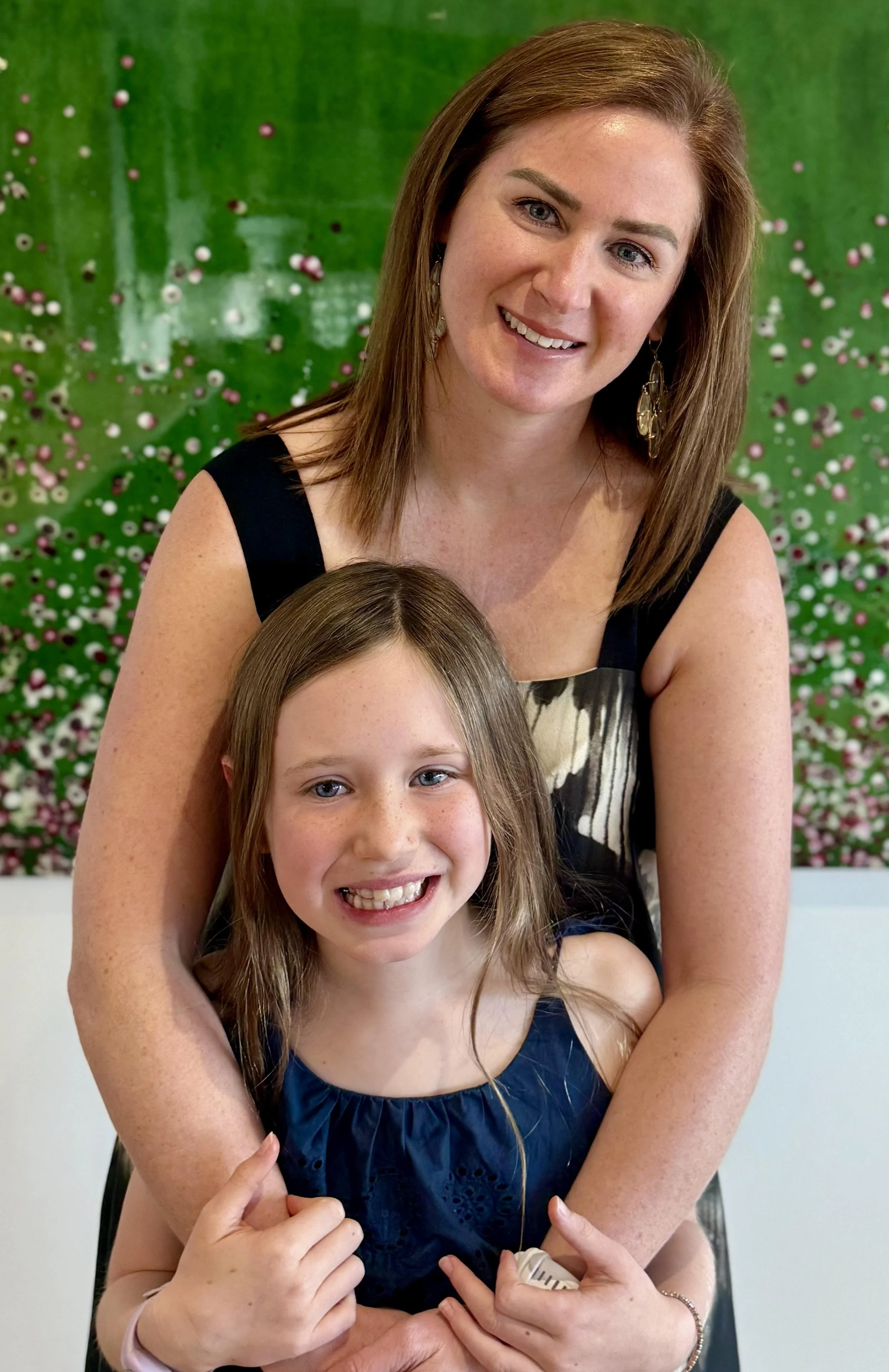 A woman with shoulder-length brown hair, wearing earrings and a black sleeveless dress, is smiling and hugging a young girl with long brown hair, who is also smiling and wearing a navy blue dress. They are standing in front of a green, colorful, blurred background with small circular patterns.