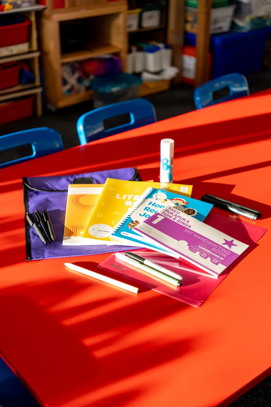 School classroom table with books, notebooks, and pens, illuminated by sunlight, with blue chairs and wooden shelves in the background.