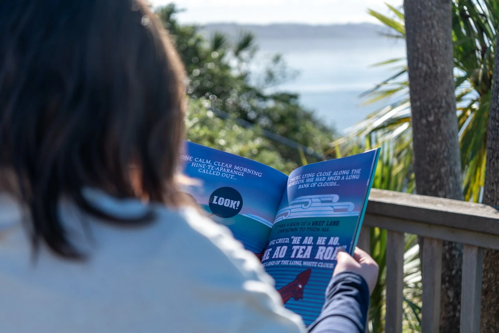 A person with dark hair reading a colorful illustrated magazine outdoors on a wooden railing, with trees and water in the background.