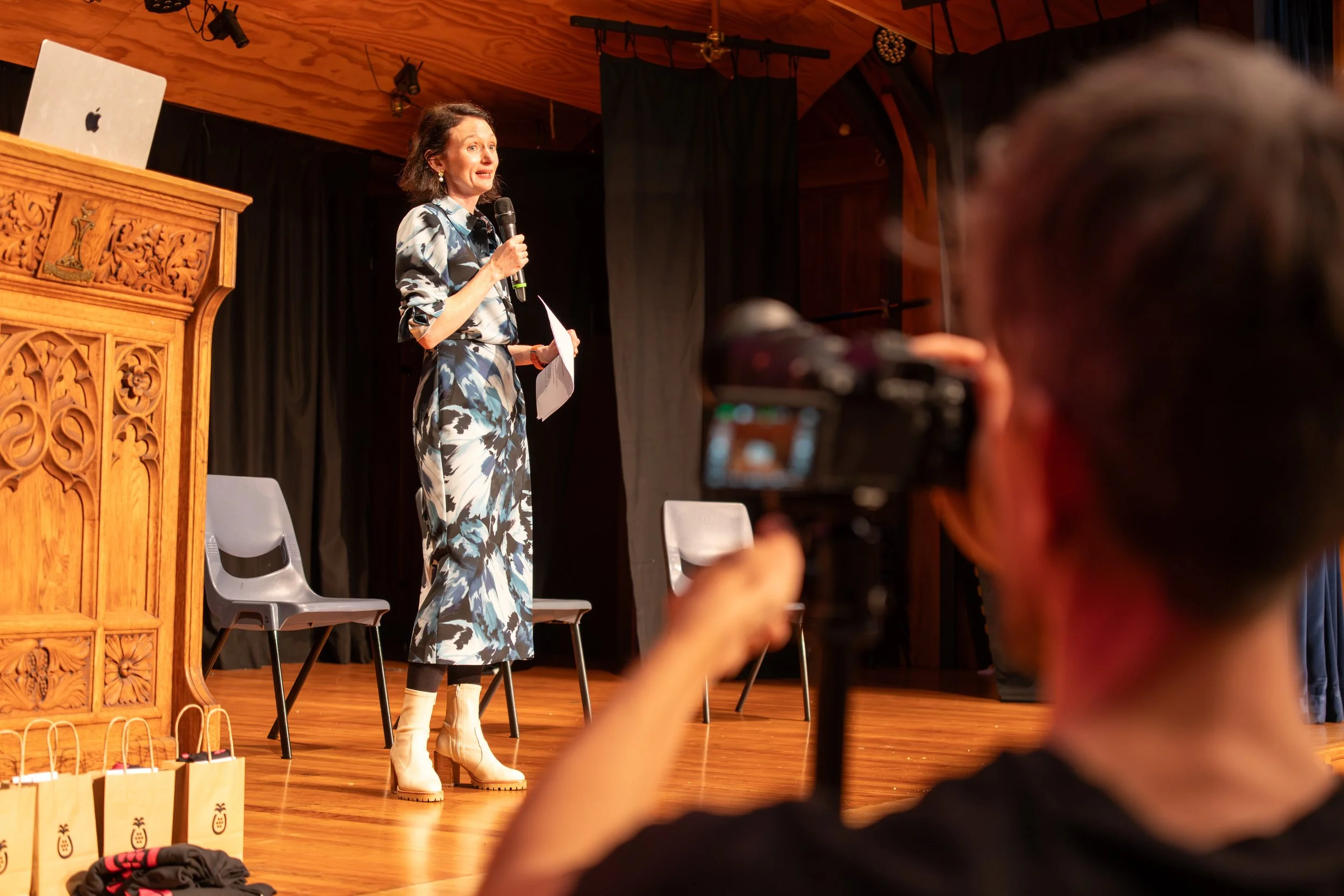 A woman standing on a stage holding a microphone, speaking to an audience. She is dressed in a blue and white patterned outfit with beige boots. There are chairs, a carved wooden podium, and gift bags on the stage. The background has black curtains and wooden walls, with stage lighting overhead.