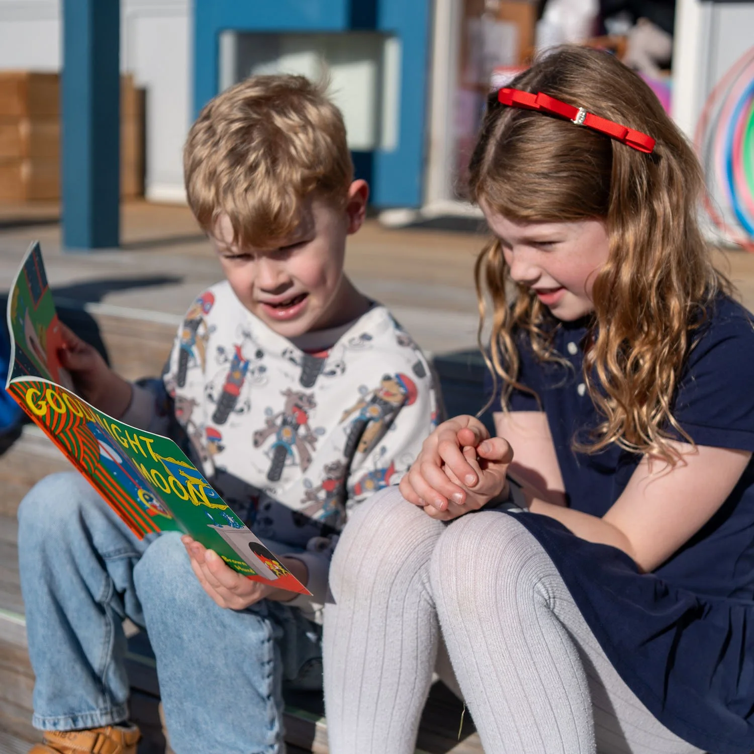 A young boy with blond hair and a young girl with brown hair and a red headband sitting outdoors, reading a colorful book together, smiling and engaging with each other.