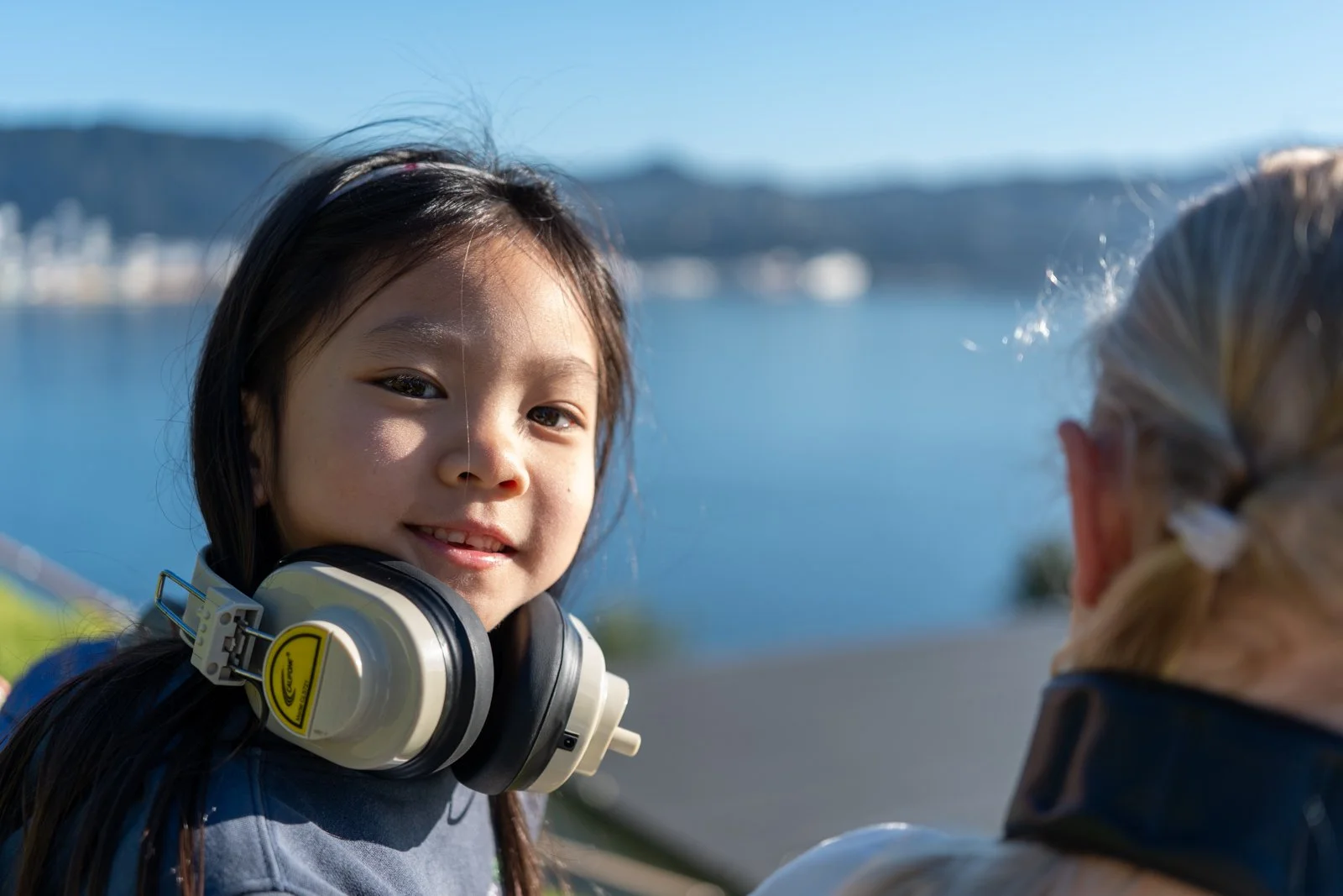A young girl with long dark hair and headphones around her neck smiling outdoors near water with a blurred person in the foreground.