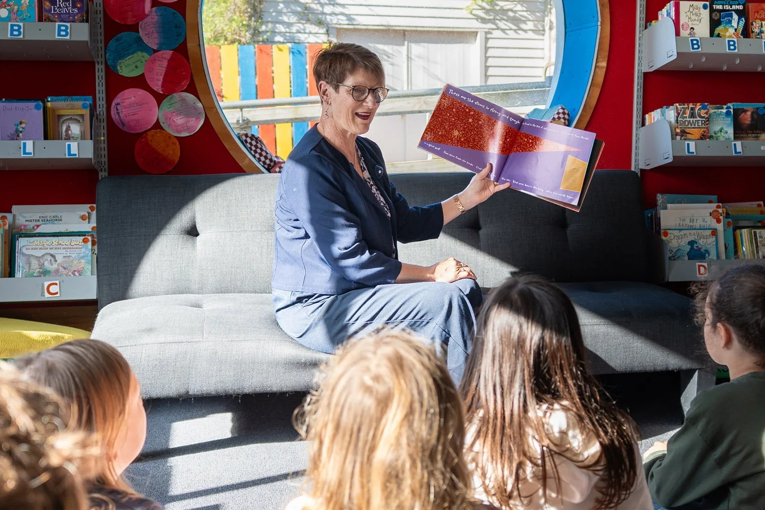 A woman reading a children's book to a group of children seated on the floor in a library or bookstore, with bookshelves and a large reflect window in the background.