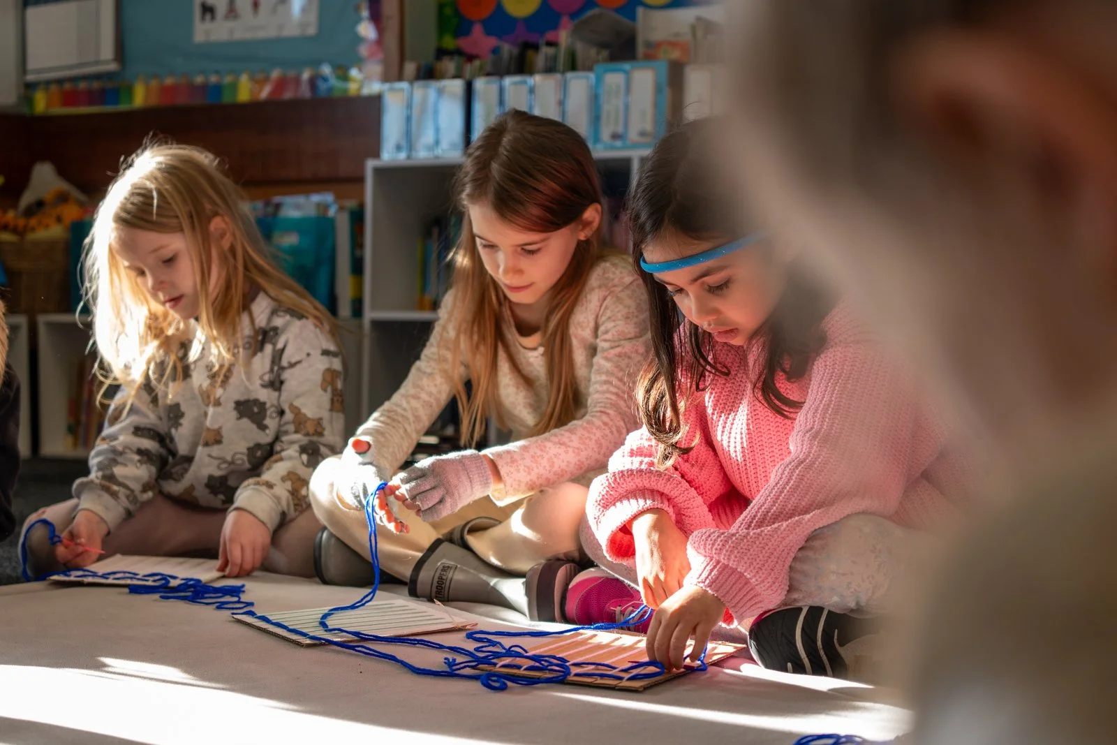 Four young girls sitting on the floor in a classroom, engaged in a craft activity with blue yarn on a white surface.