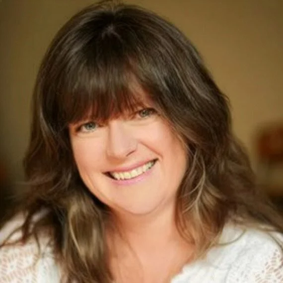 A woman with long brown hair and bangs, smiling with visible teeth, wearing a white top, against a warm background.