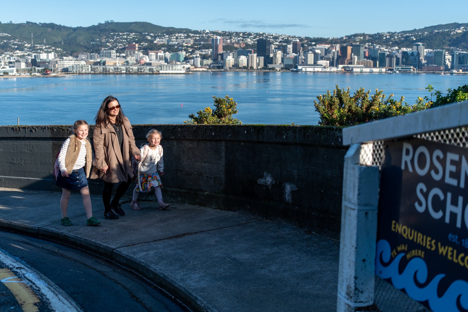 A woman walking hand-in-hand with two young girls along a waterfront promenade with a city skyline and hills in the background.