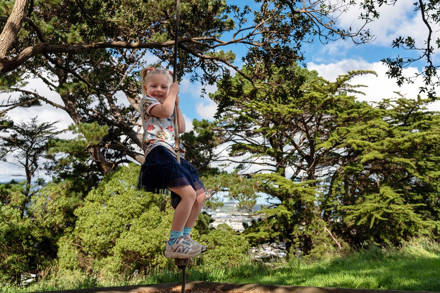 A young girl smiling and playing on a rope swing outdoors surrounded by green trees and grass, with a partly cloudy sky in the background.