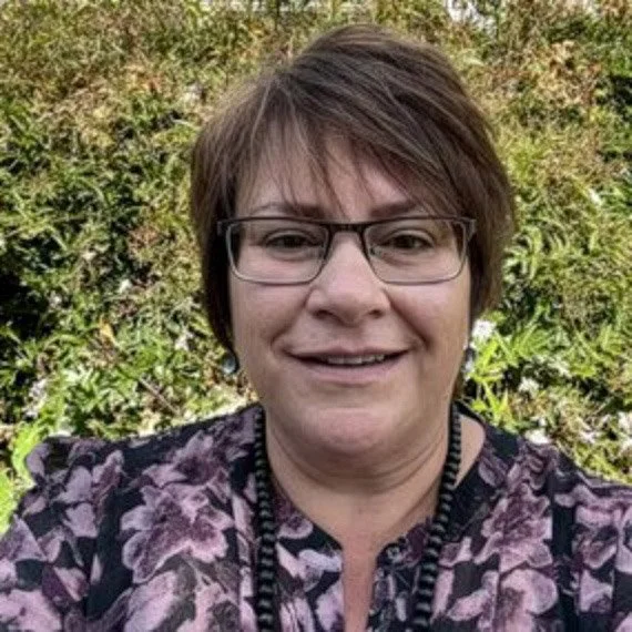 A woman with short brown hair and glasses smiling outdoors in front of green foliage.