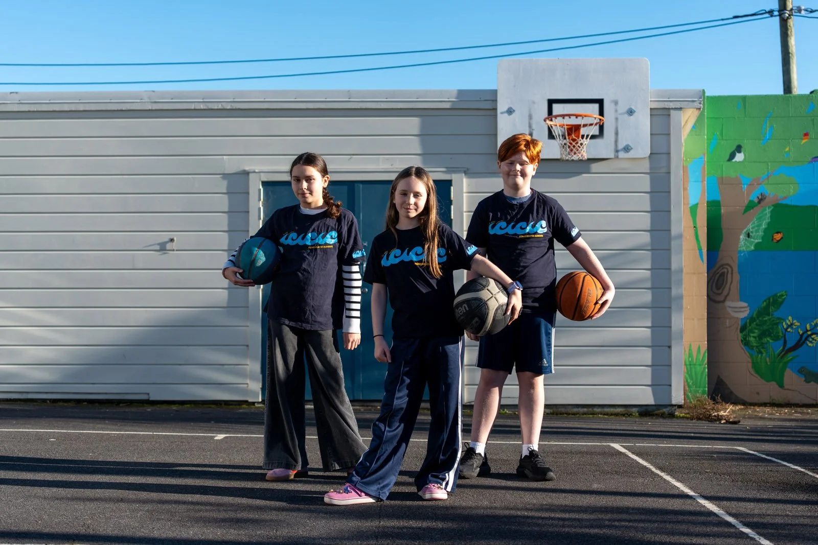 Three children standing on a basketball court in front of a wall with a mural, each holding a basketball, dressed in sports uniforms.