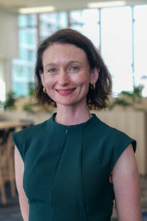 A woman with shoulder-length dark hair, wearing a teal sleeveless top, smiling in an office setting with large windows and a modern interior.