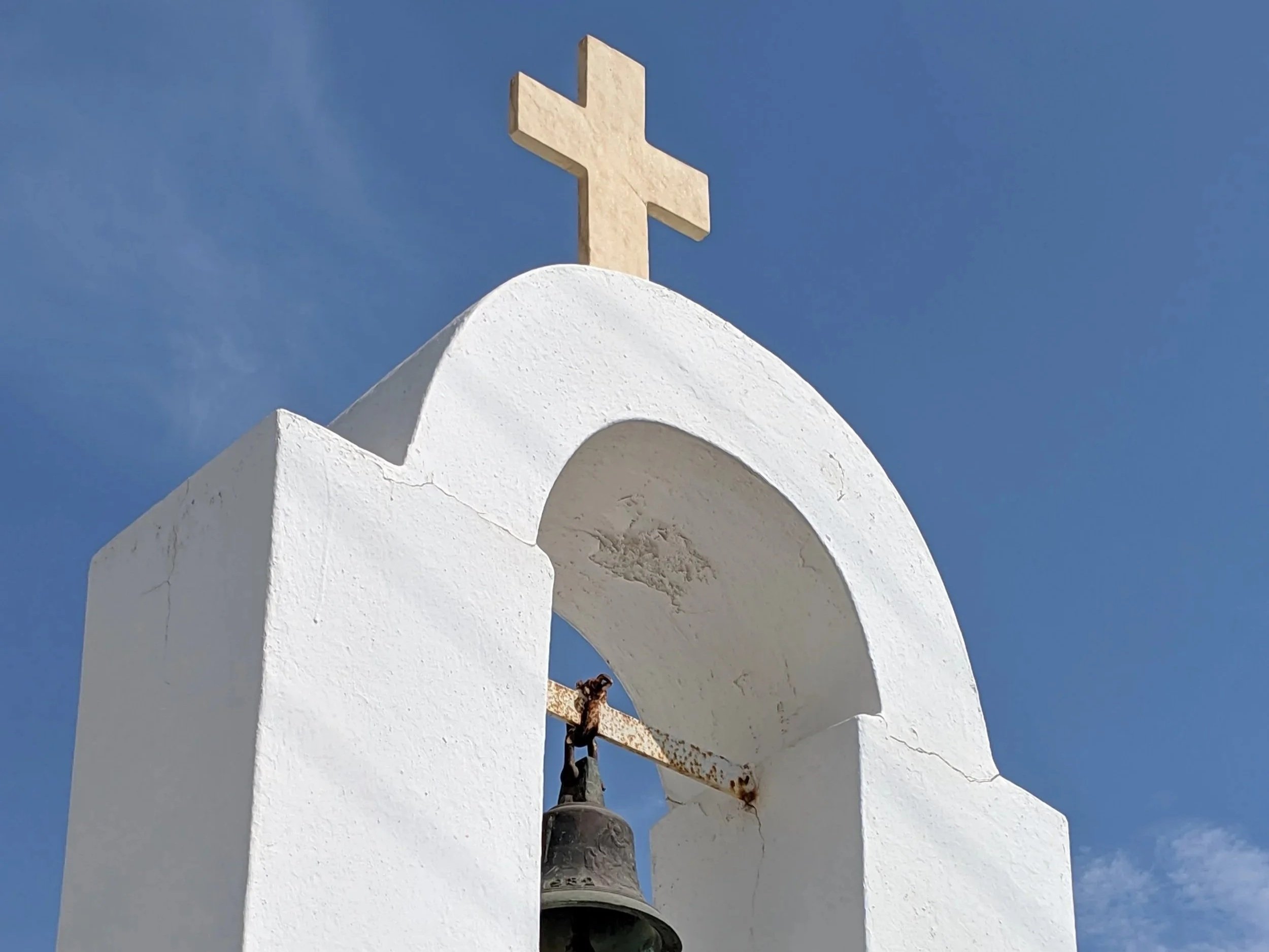 A white church steeple with a cross at the top, against a clear blue sky, with a rusty bell and a chain hanging from the structure.