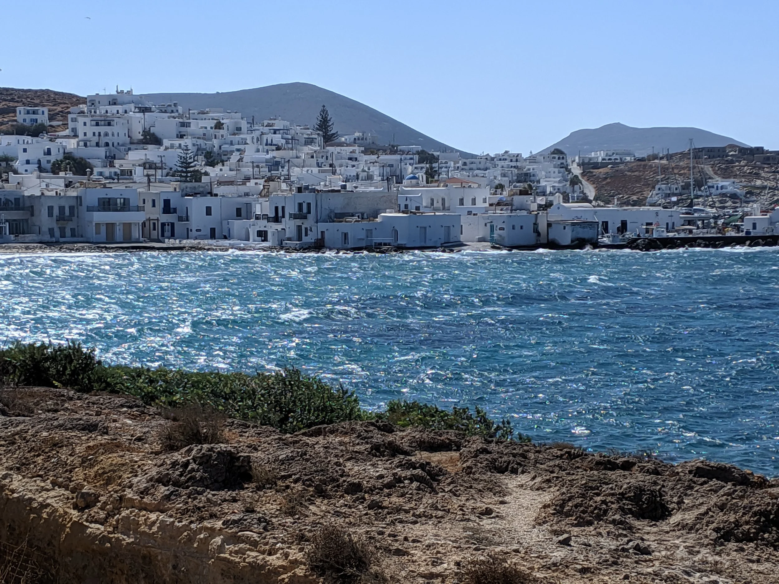A seaside town with white buildings on a hillside next to the blue ocean, with mountains in the background and a clear sky.