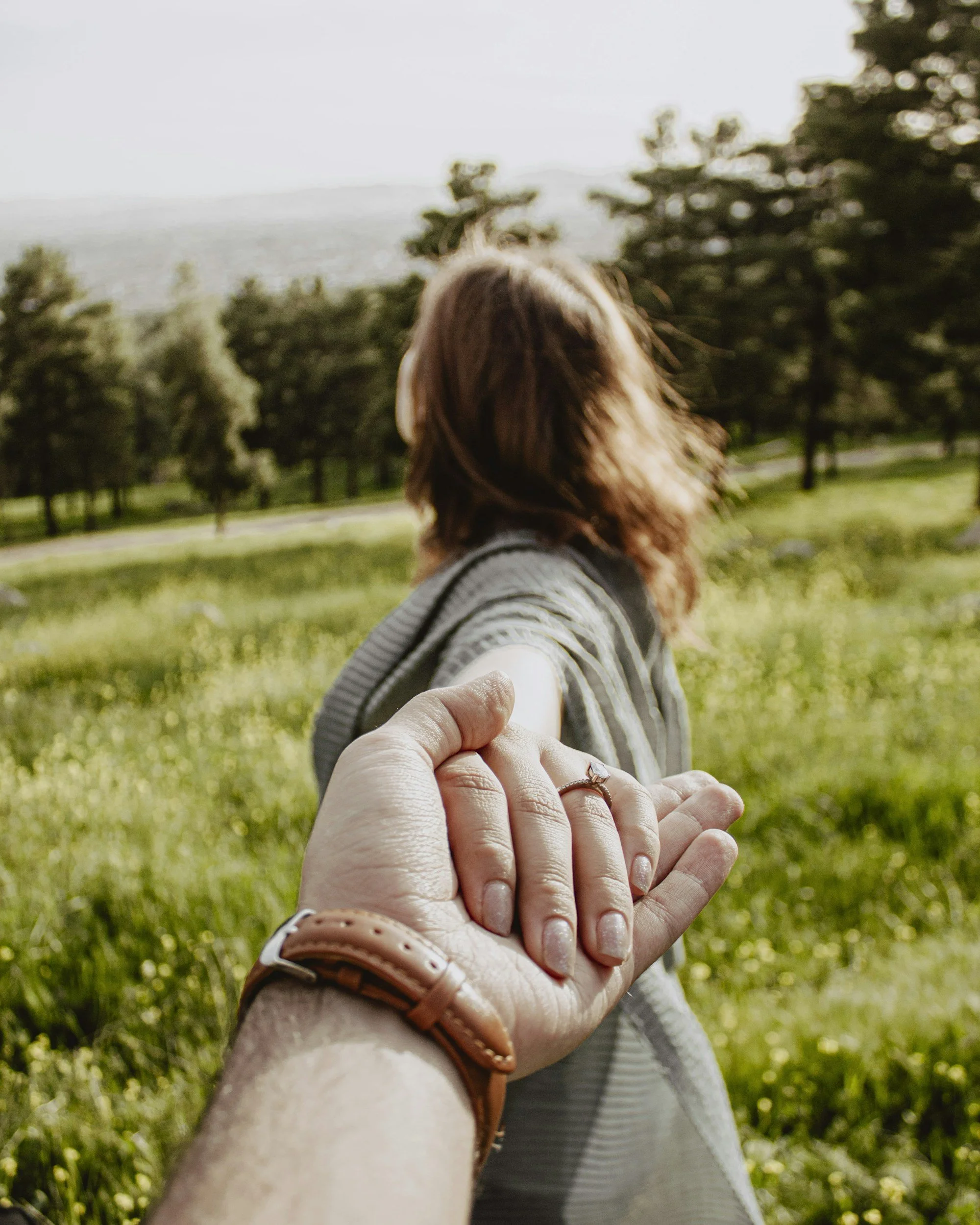 A person with long reddish-brown hair is reaching out their hand to a child, who is holding their hand while walking outdoors in a grassy area with trees and hills in the background.