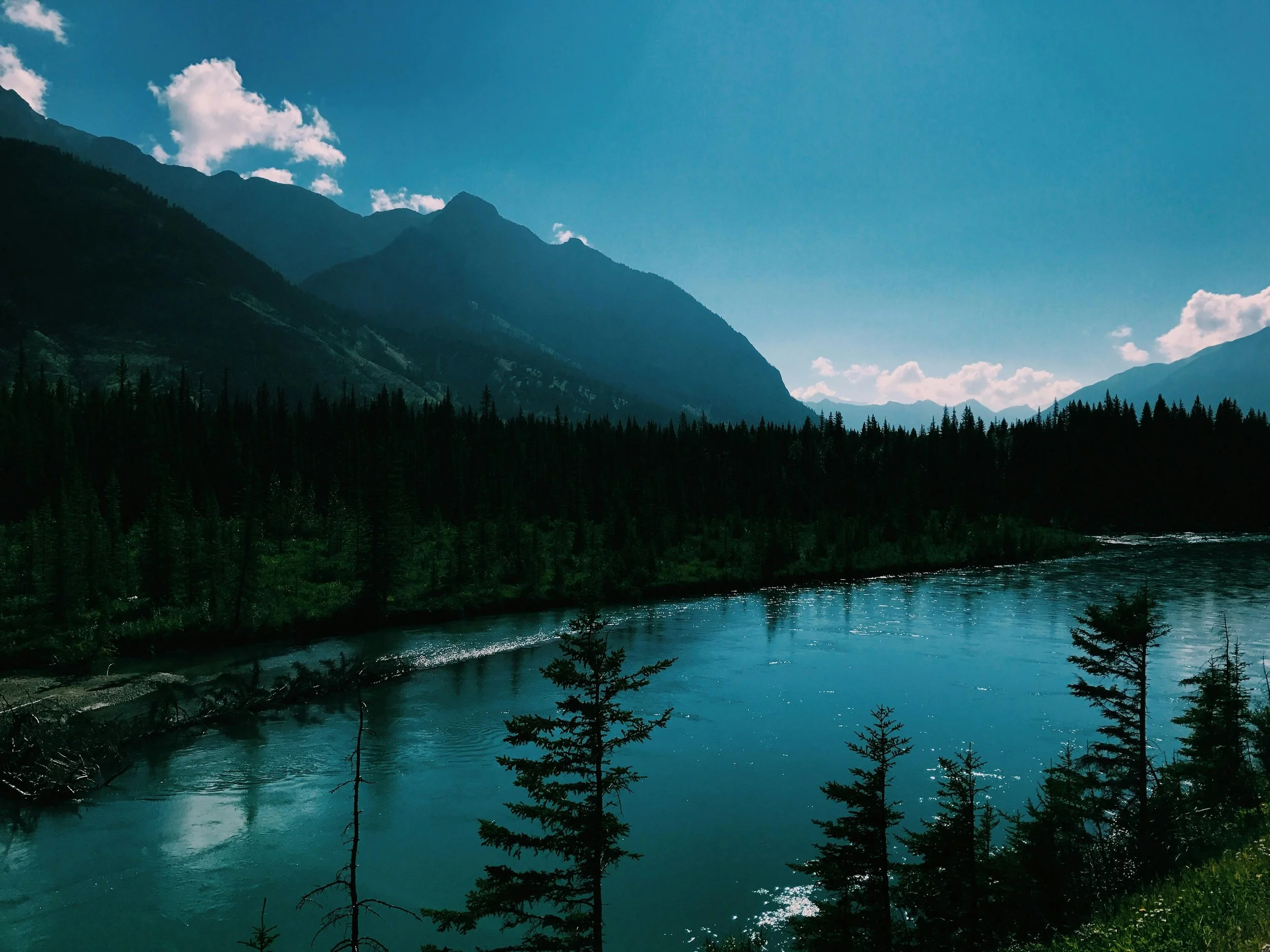 Scenic view of a mountain range with lush green forests, a river in the foreground, and clouds scattered in a clear blue sky.