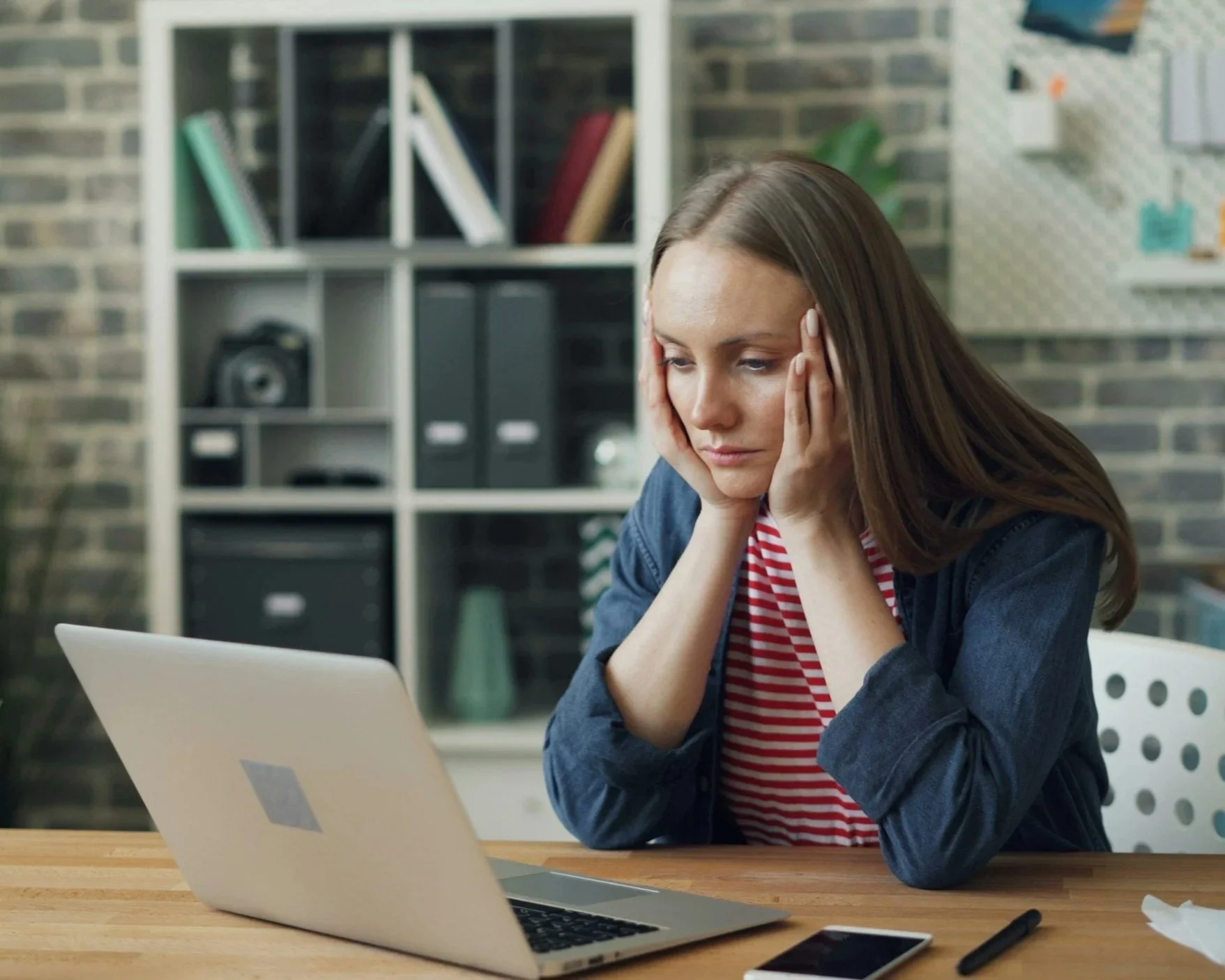 `Woman sitting at a desk with a laptop, looking downward with a thoughtful expression, reflecting the self-doubt and second-guessing common in imposter syndrome`