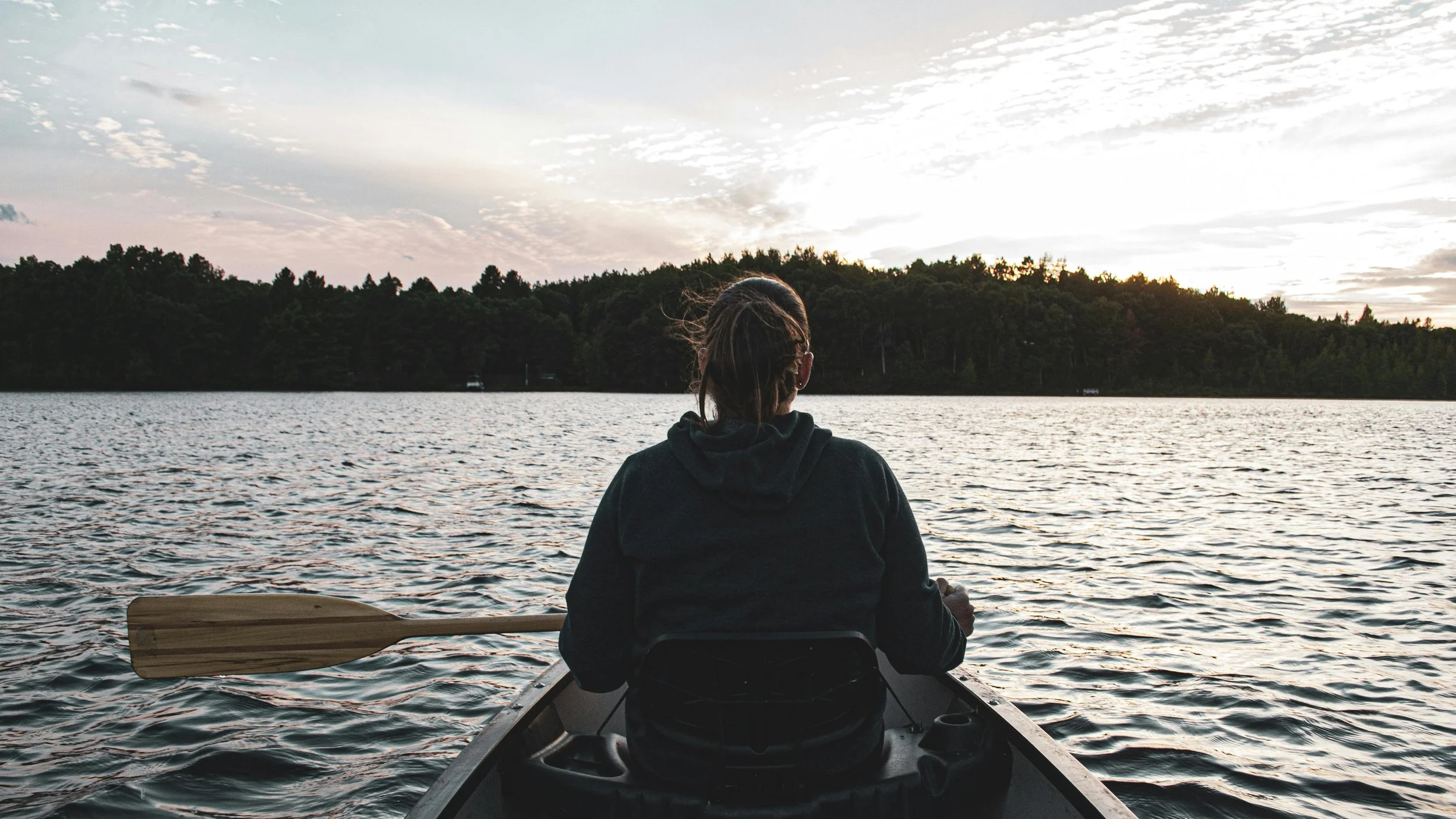 A person with braided hair, wearing a dark hoodie, sitting in a small boat on a lake, facing away, with a wooden paddle resting inside the boat, during sunset with trees across the water.