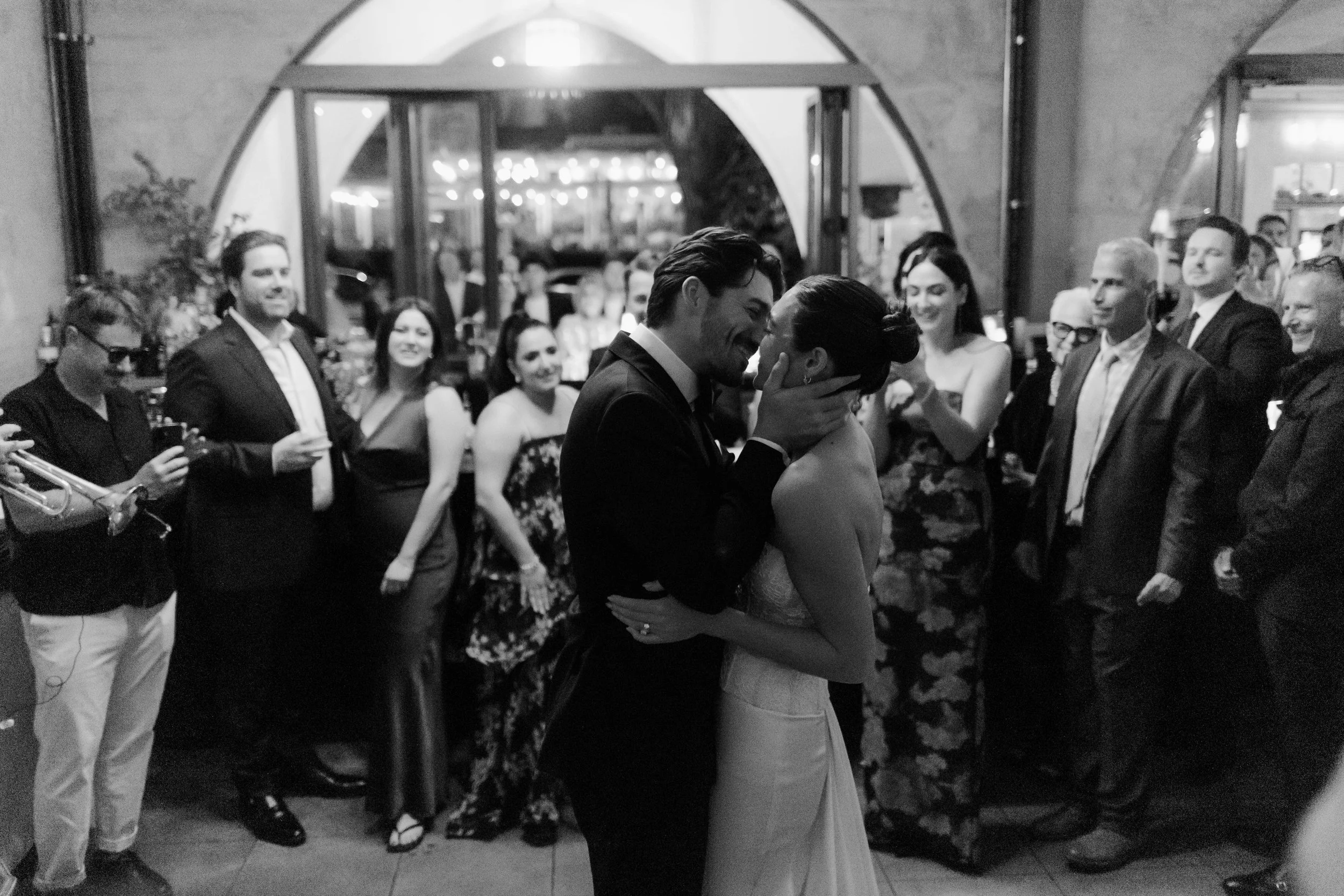 A black-and-white photo of a couple sharing a kiss and dancing at a wedding reception, surrounded by guests who are smiling and watching them.
