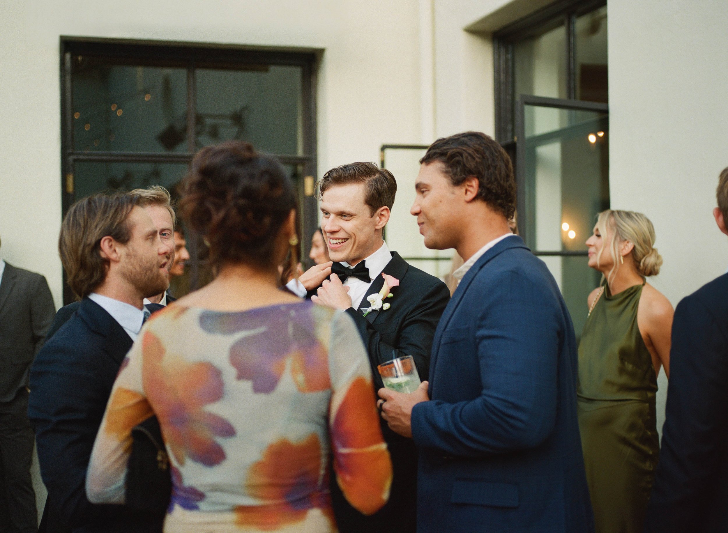 People at a social gathering engaged in conversation, with men in tuxedos and women in colorful dresses inside a room with large windows.