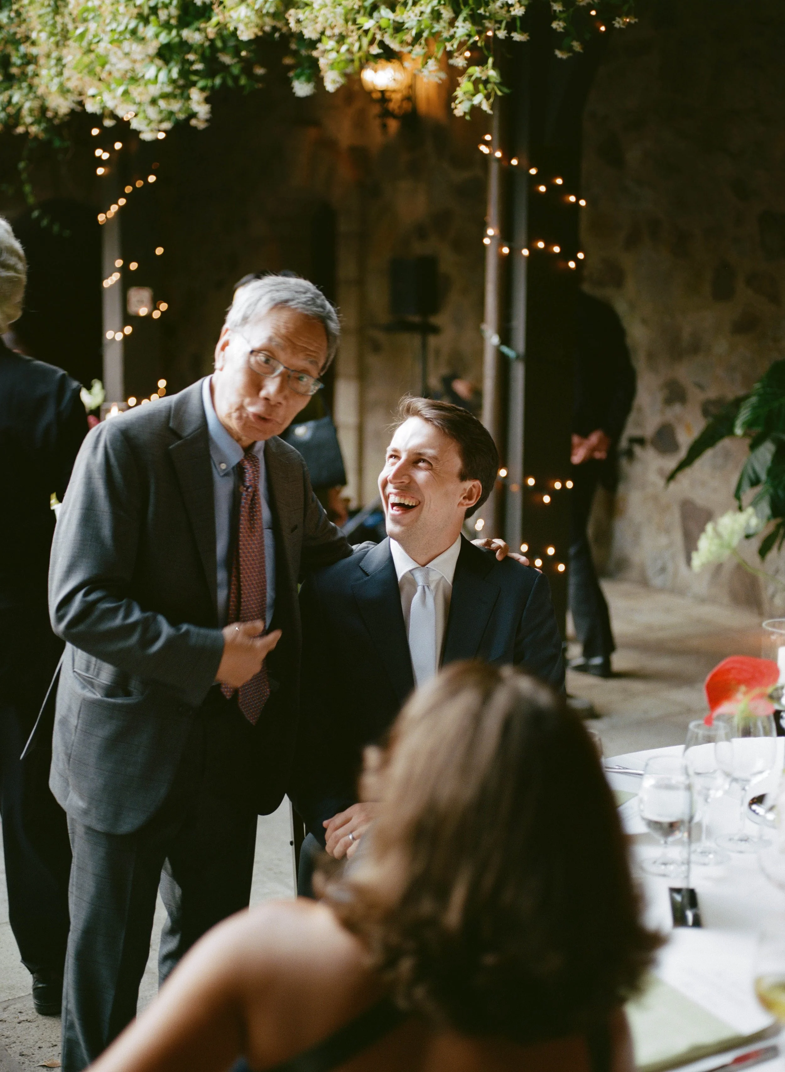 Two men conversing at a formal event, with decorated string lights hanging in the background, and a table with glasses and plates in the foreground.