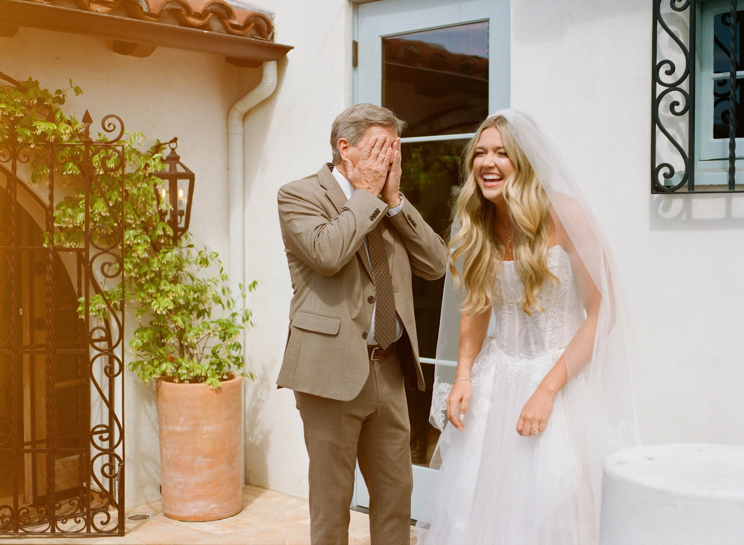 A man in a suit covering his face with his hands and a woman in a white dress laughing in front of a door. They appear to be celebrating a wedding.