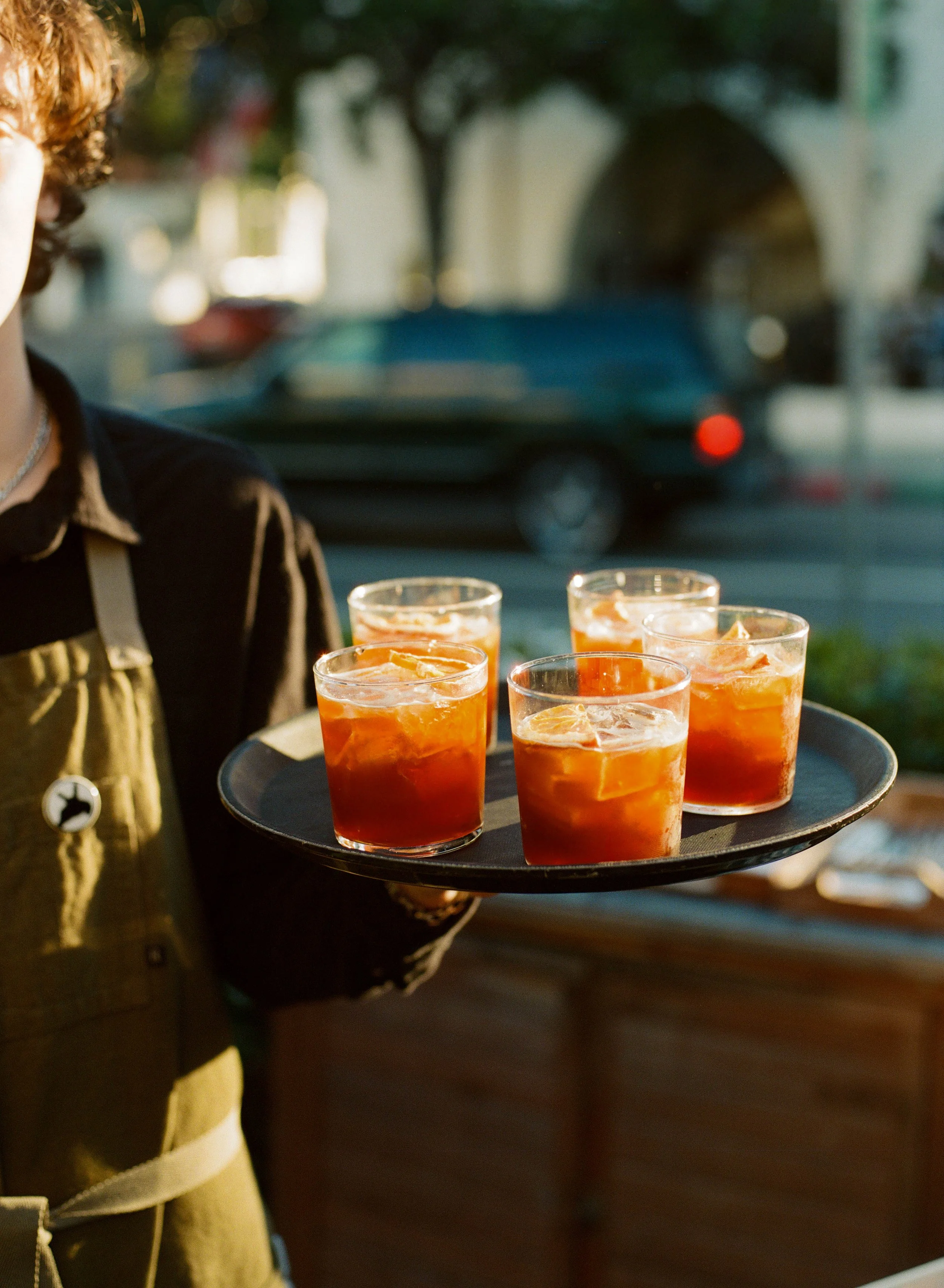 Person holding a tray of four glasses filled with orange-colored beverages, garnished with slices of citrus fruit, outdoors during daylight.
