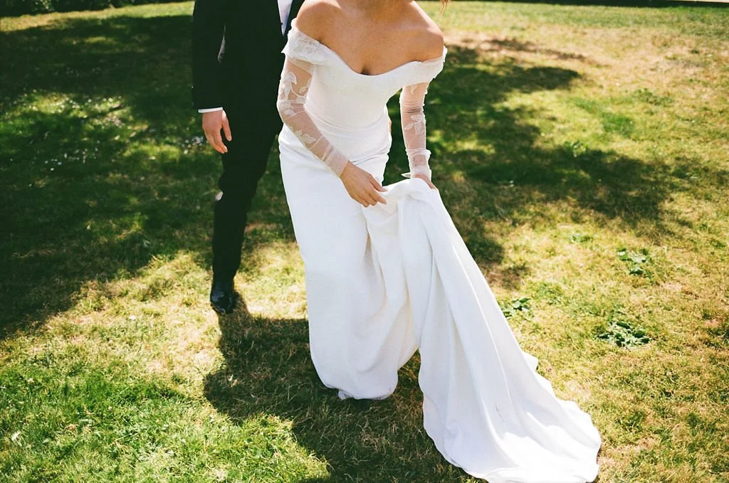 A bride in a white wedding dress with lace sleeves holding up her gown, standing on grass during the daytime. A groom in a black suit stands behind her.