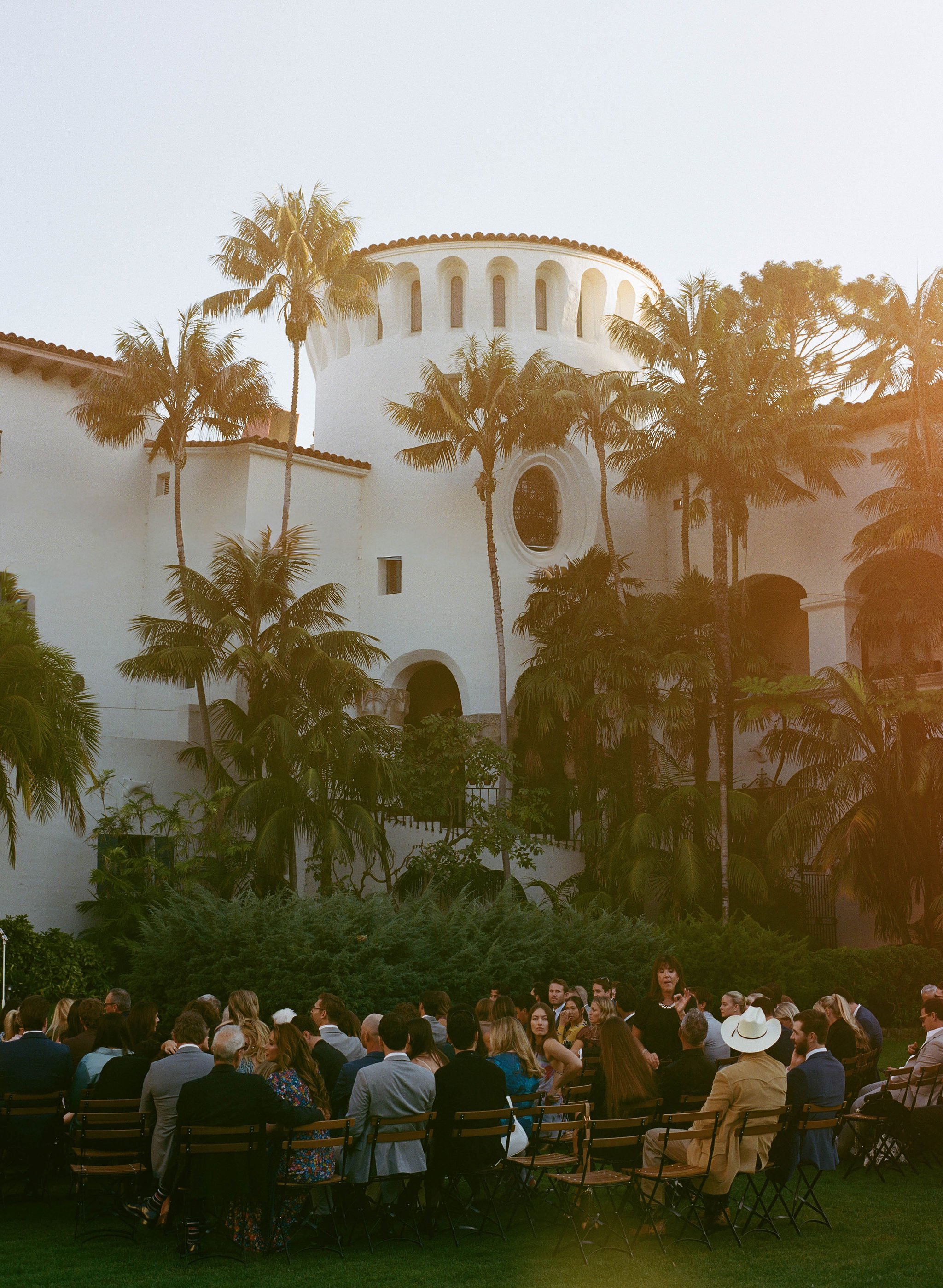 Outdoor gathering with numerous people seated in chairs, surrounded by lush greenery and tall palm trees, with a large white building featuring a rounded tower and arched windows in the background. The scene is illuminated by warm sunlight, suggestin